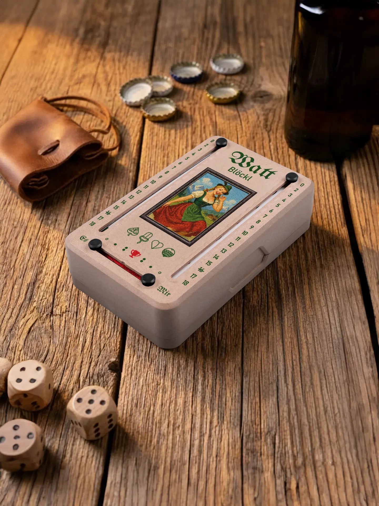 Wattblöckl in Holzoptik mit schwarzen Pins und Alpenmadl-Motiv. Platziert auf Holztisch mit Würfeln und Kronkorken.