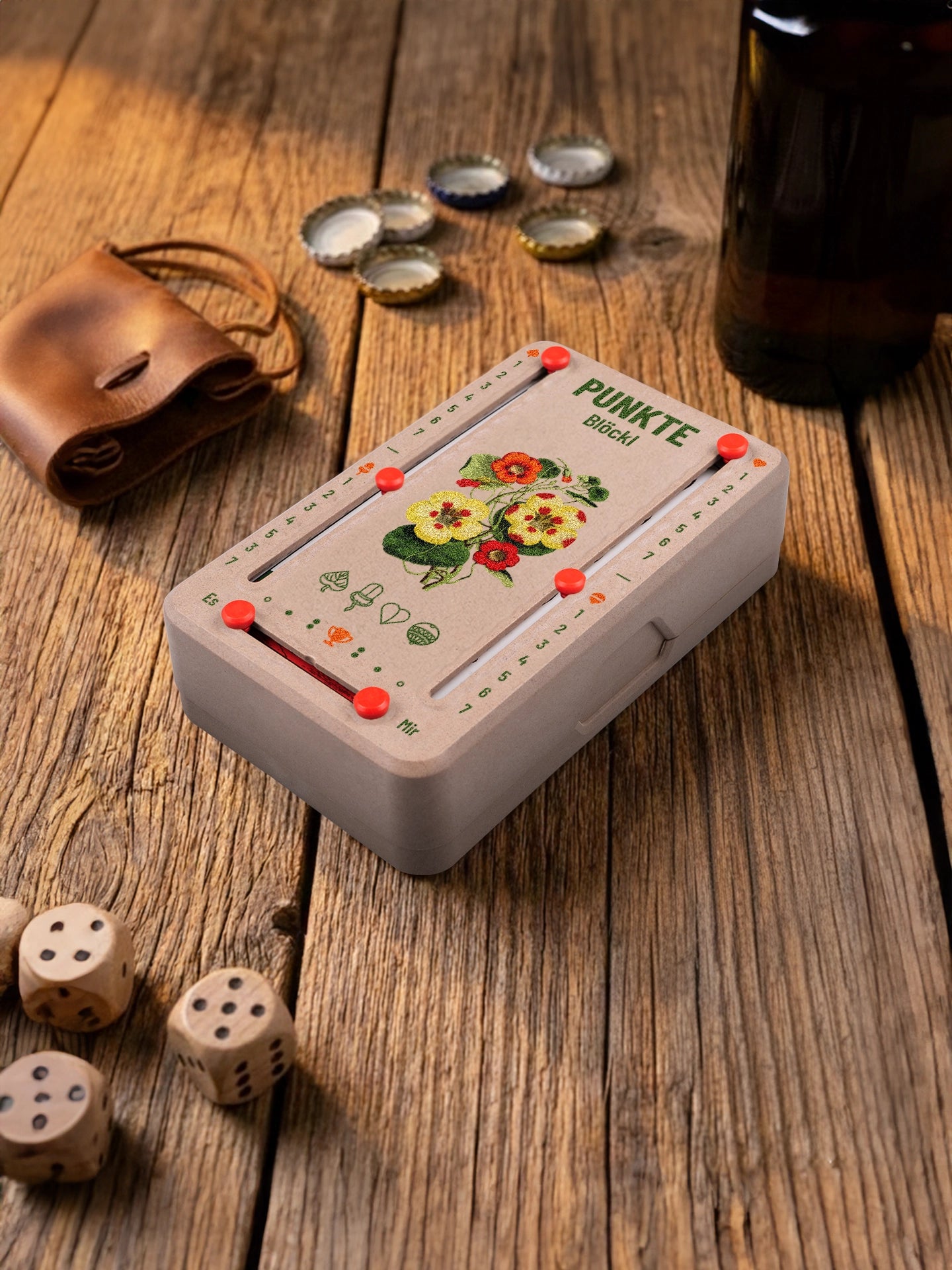 Punkteblöckl in Holzoptik mit roten Pins und Bluetenblatt-Motiv. Platziert auf Holztisch mit Würfeln und Kronkorken.