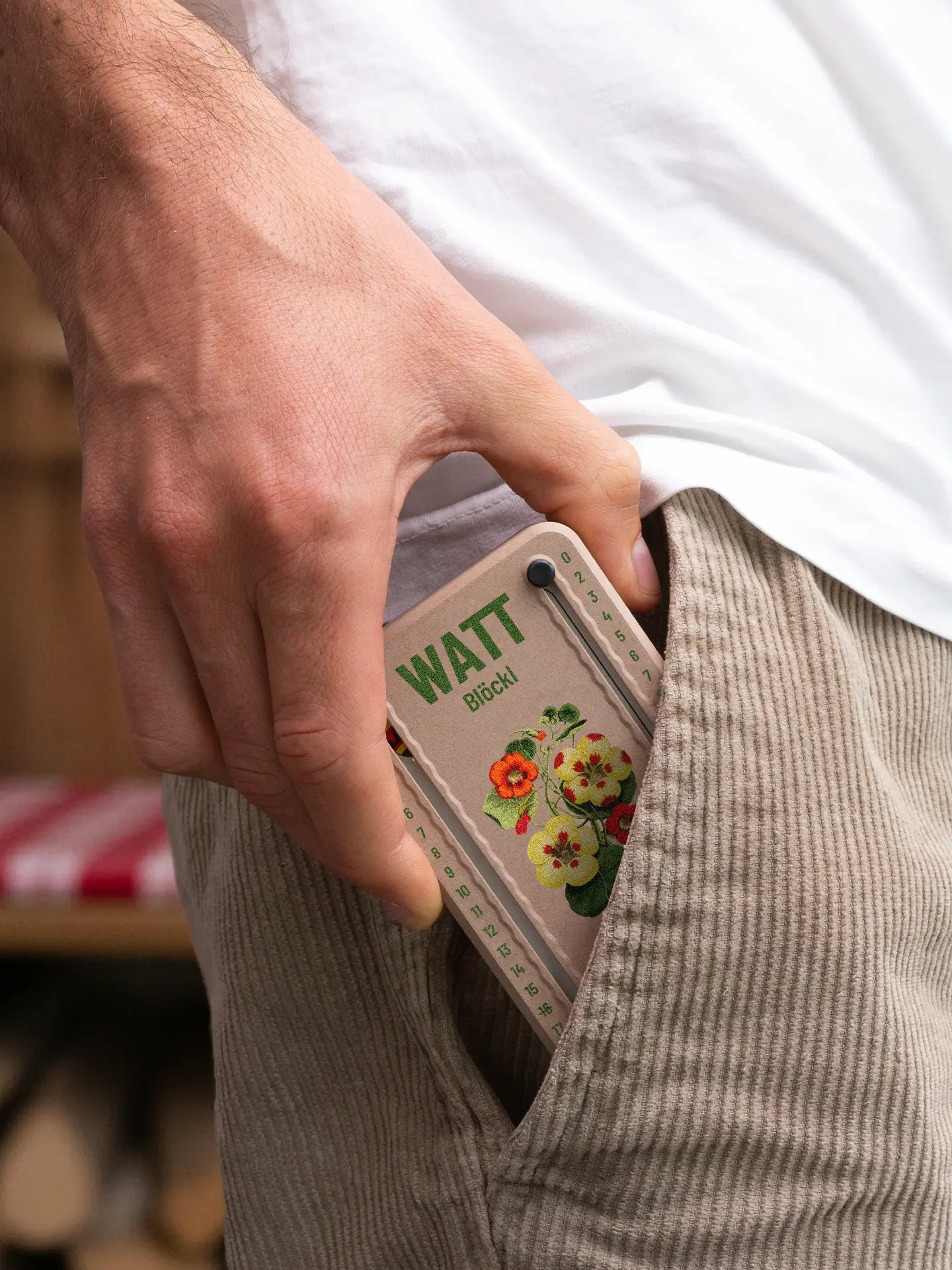 Wattblöckl in Holzoptik mit roten Pins und Bluetenblatt-Motiv wird aus der Hosentasche gezogen.