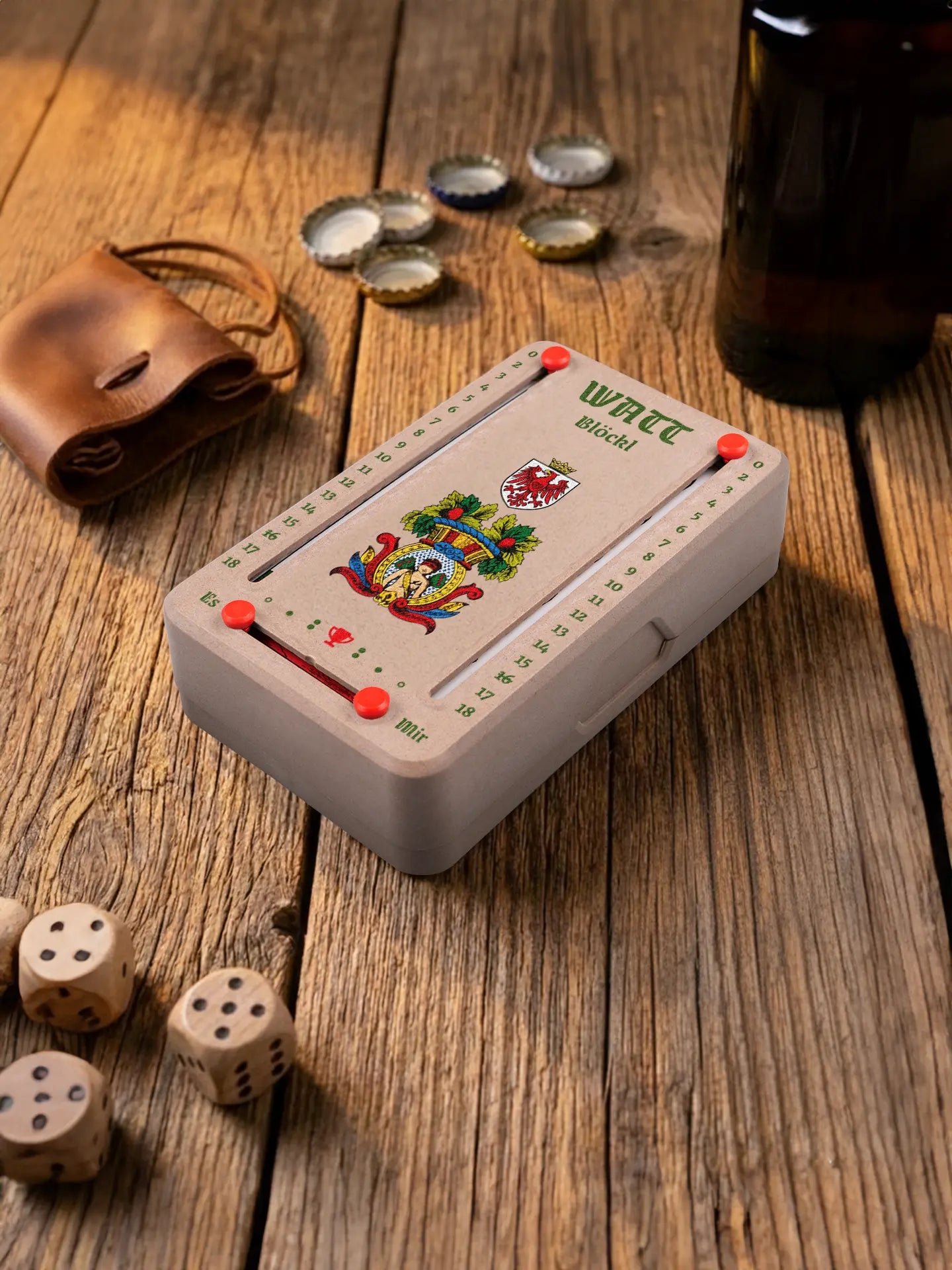 Wattblöckl in Holzoptik mit roten Pins und HerzAss-Motiv. Platziert auf Holztisch mit Würfeln und Kronkorken.