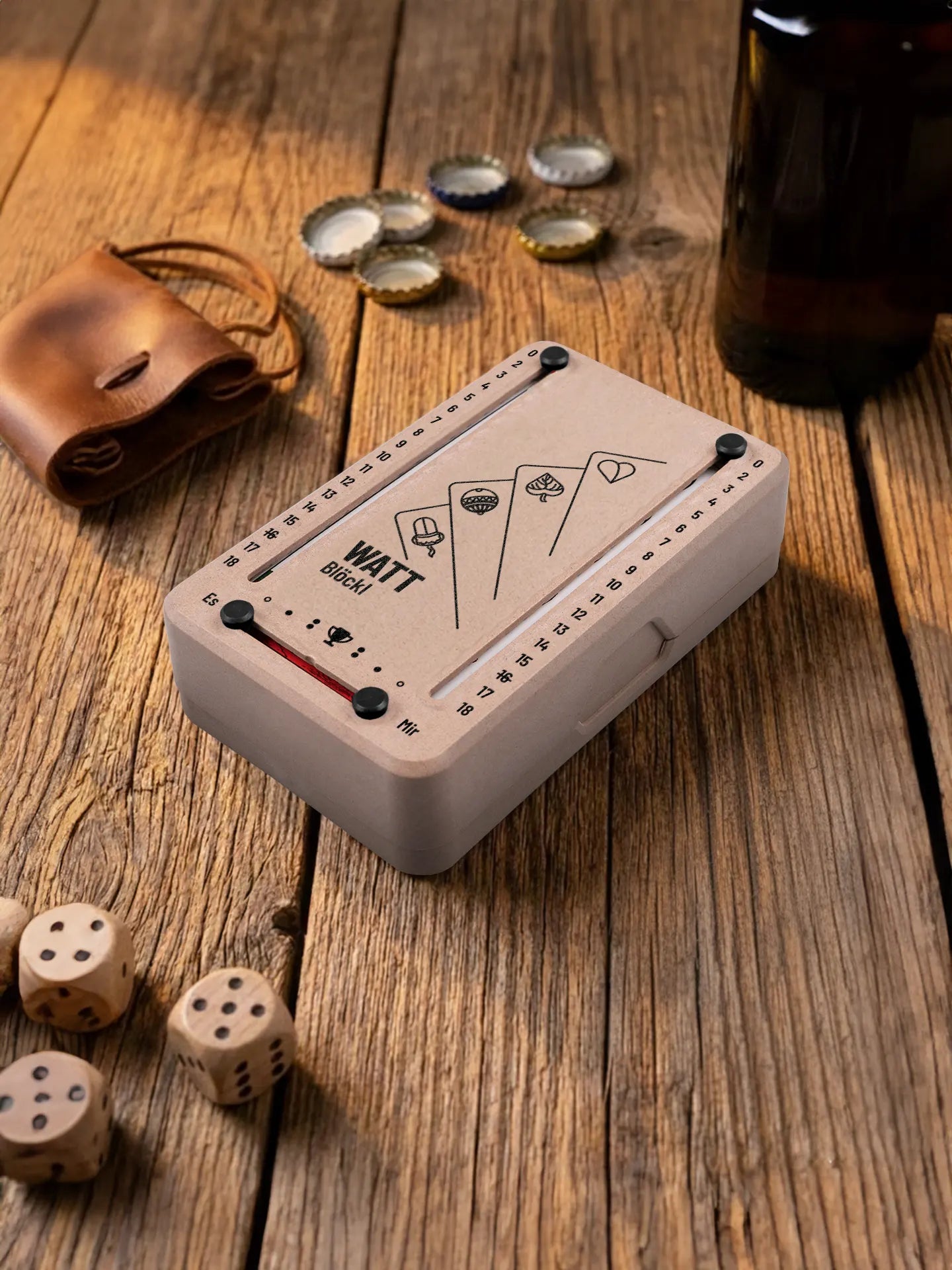 Wattblöckl in Holzoptik mit schwarzen Pins und Karten-Motiv. Platziert auf Holztisch mit Würfeln und Kronkorken.