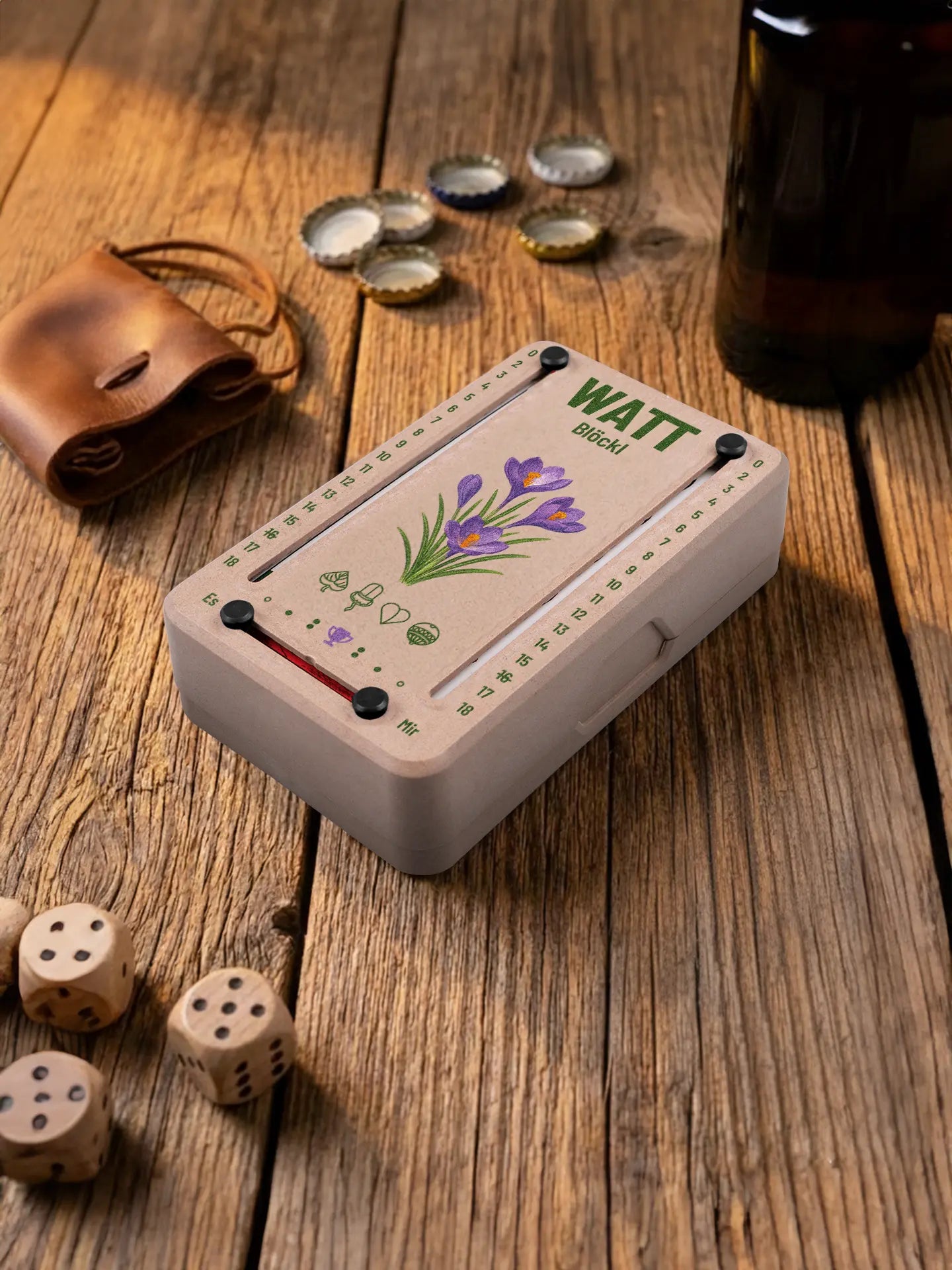 Wattblöckl in Holzoptik mit schwarzen Pins und Krokus-Motiv. Platziert auf Holztisch mit Würfeln und Kronkorken.