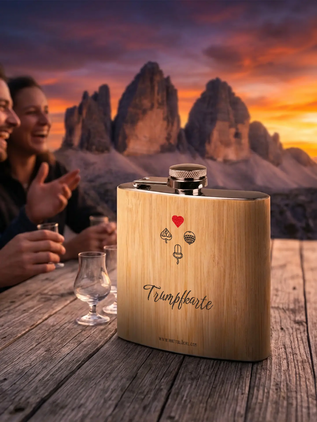 Flachmann bei Sonnenuntergang, leicht seitlich fotografiert und auf einem Holztisch in Szene gesetzt. Im Hintergrund eine stimmungsvolle Berglandschaft mit Personen.