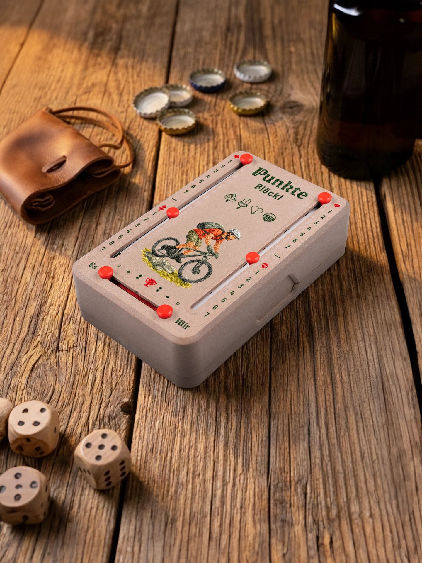 Punkteblöckl in Holzoptik mit roten Pins und Mountainbiker-Motiv. Platziert auf Holztisch mit Würfeln und Kronkorken.