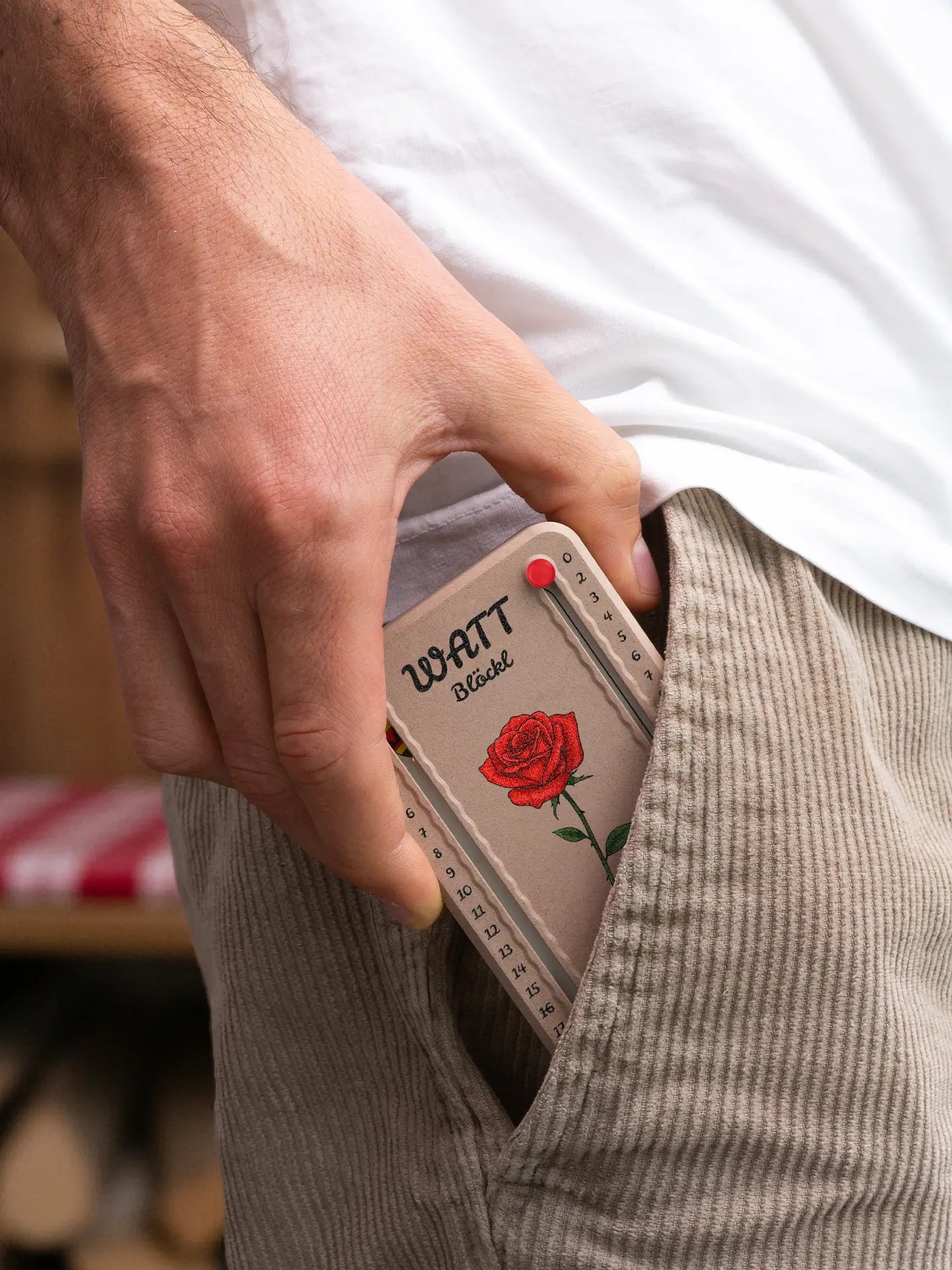 Wattblöckl in Holzoptik mit roten Pins und Rose-Motiv wird aus der Hosentasche gezogen.