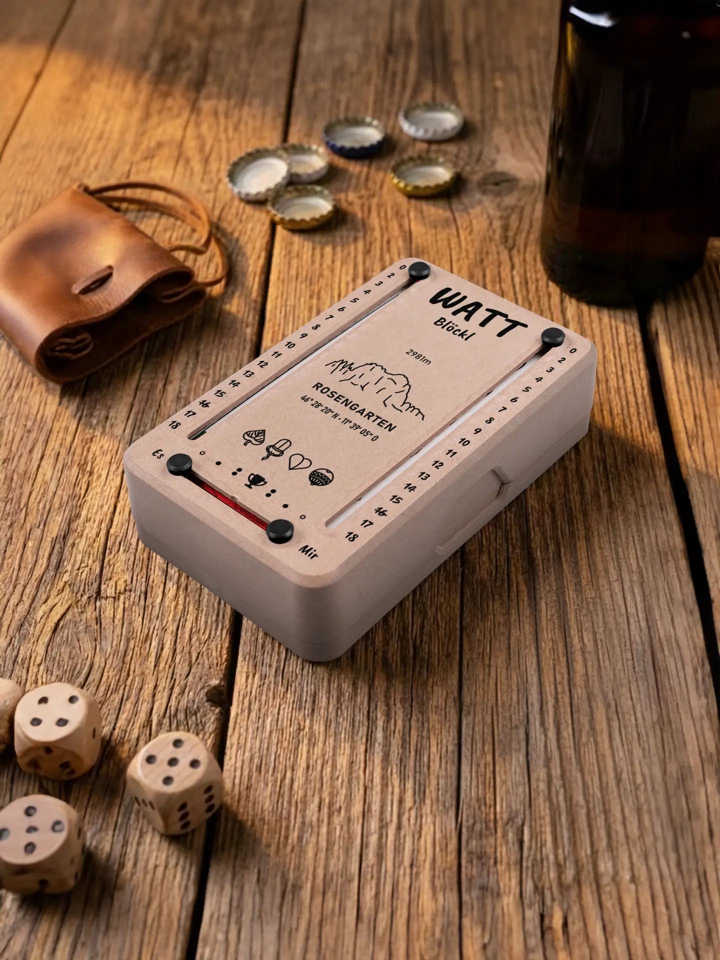 Wattblöckl in Holzoptik mit schwarzen Pins und Schlern-Motiv. Platziert auf Holztisch mit Würfeln und Kronkorken.