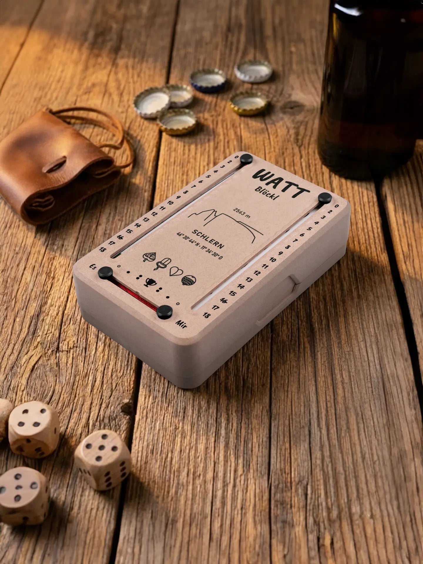 Wattblöckl in Holzoptik mit schwarzen Pins und Schlern-Motiv. Platziert auf Holztisch mit Würfeln und Kronkorken.