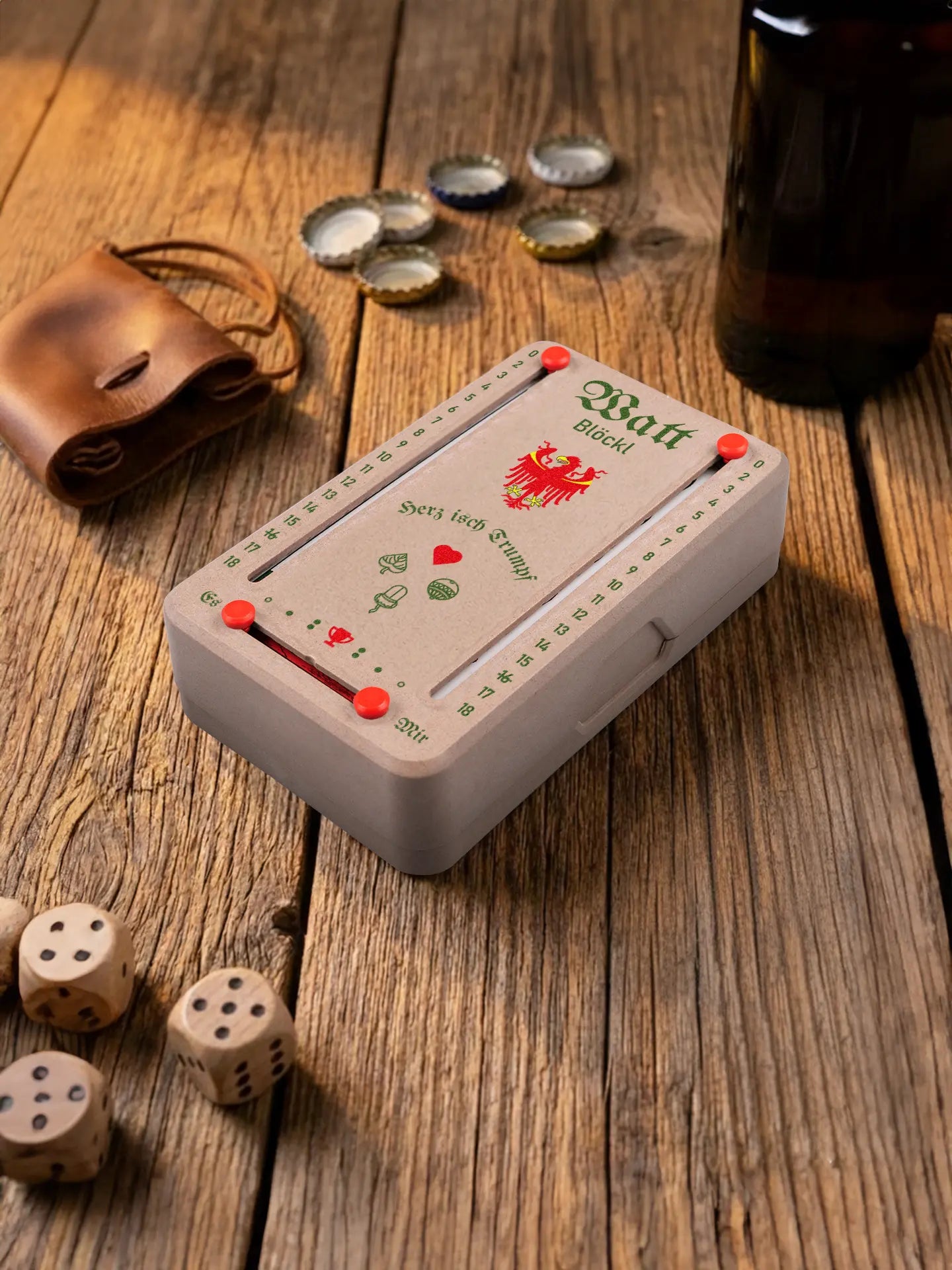 Wattblöckl in Holzoptik mit roten Pins und Suedtirol-Motiv. Platziert auf Holztisch mit Würfeln und Kronkorken.