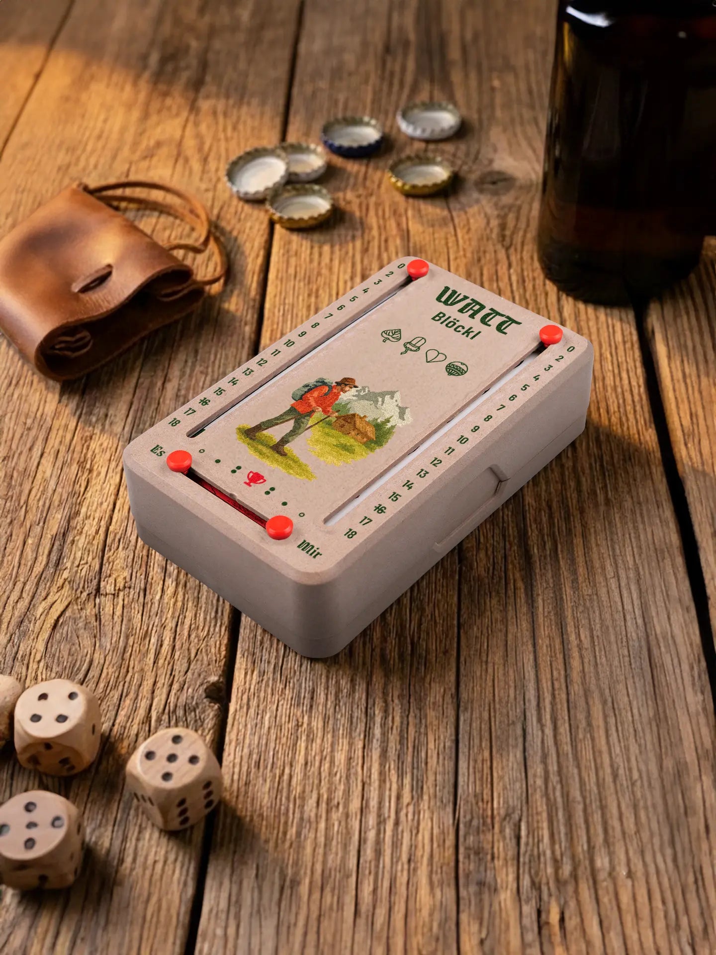 Wattblöckl in Holzoptik mit roten Pins und Wanderer-Motiv. Platziert auf Holztisch mit Würfeln und Kronkorken.